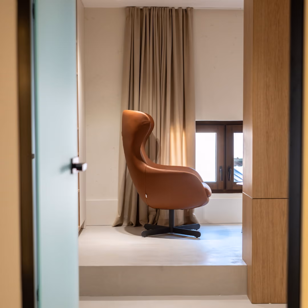 Modern office interior with wooden panel divider, brown leather armchairs, minimalist desk with laptop, lamp, and decorative head sculpture.