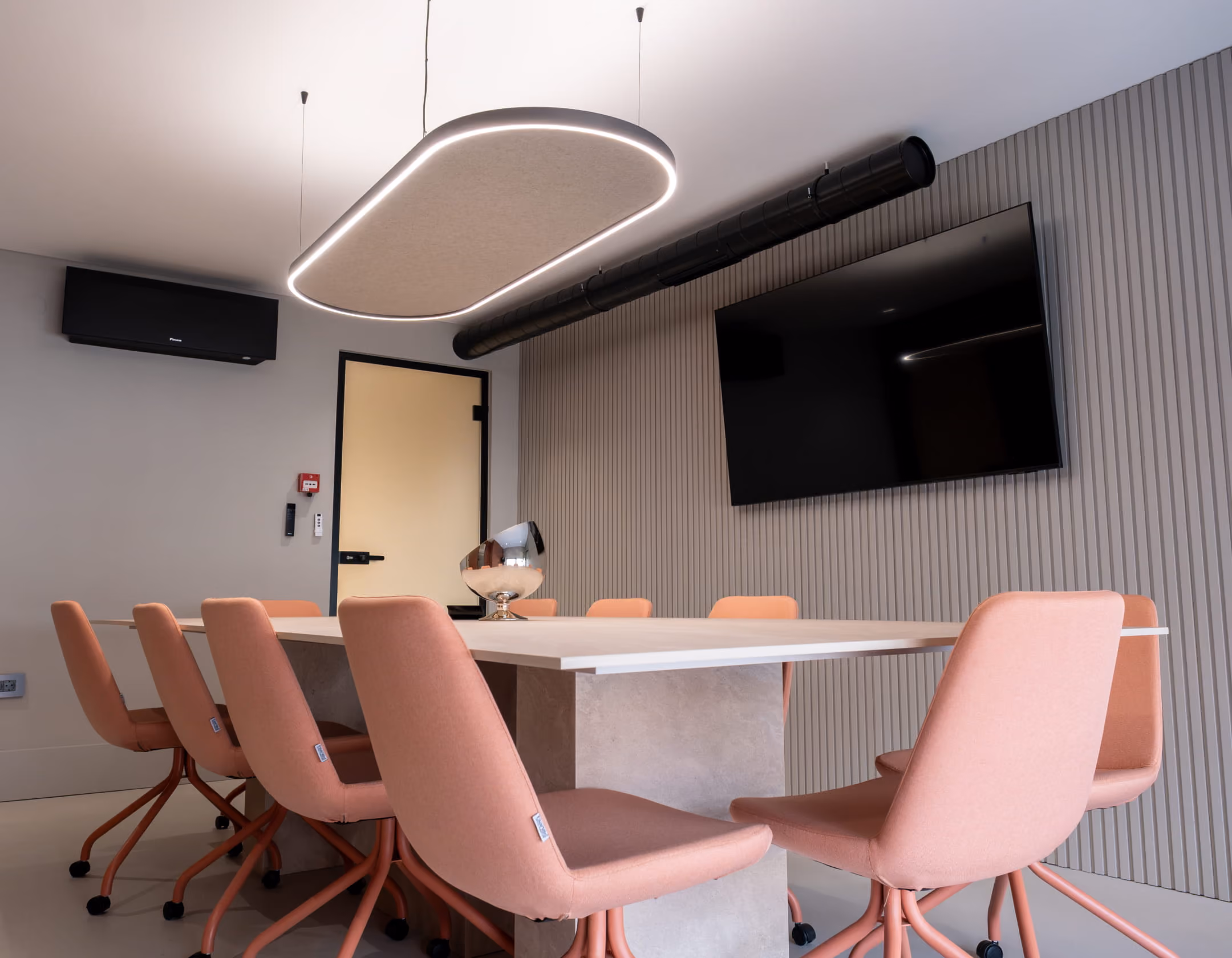 Modern conference room with a black table, eight beige swivel chairs, a wall-mounted TV, and a ring-shaped pendant light.