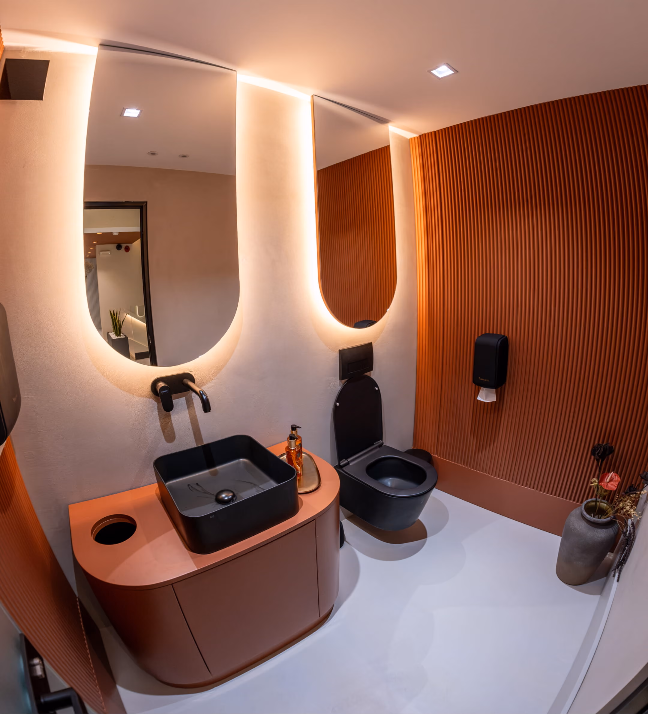 Modern bathroom with brown walls, black sink and toilet, and two vertical backlit mirrors on a gray stone wall.