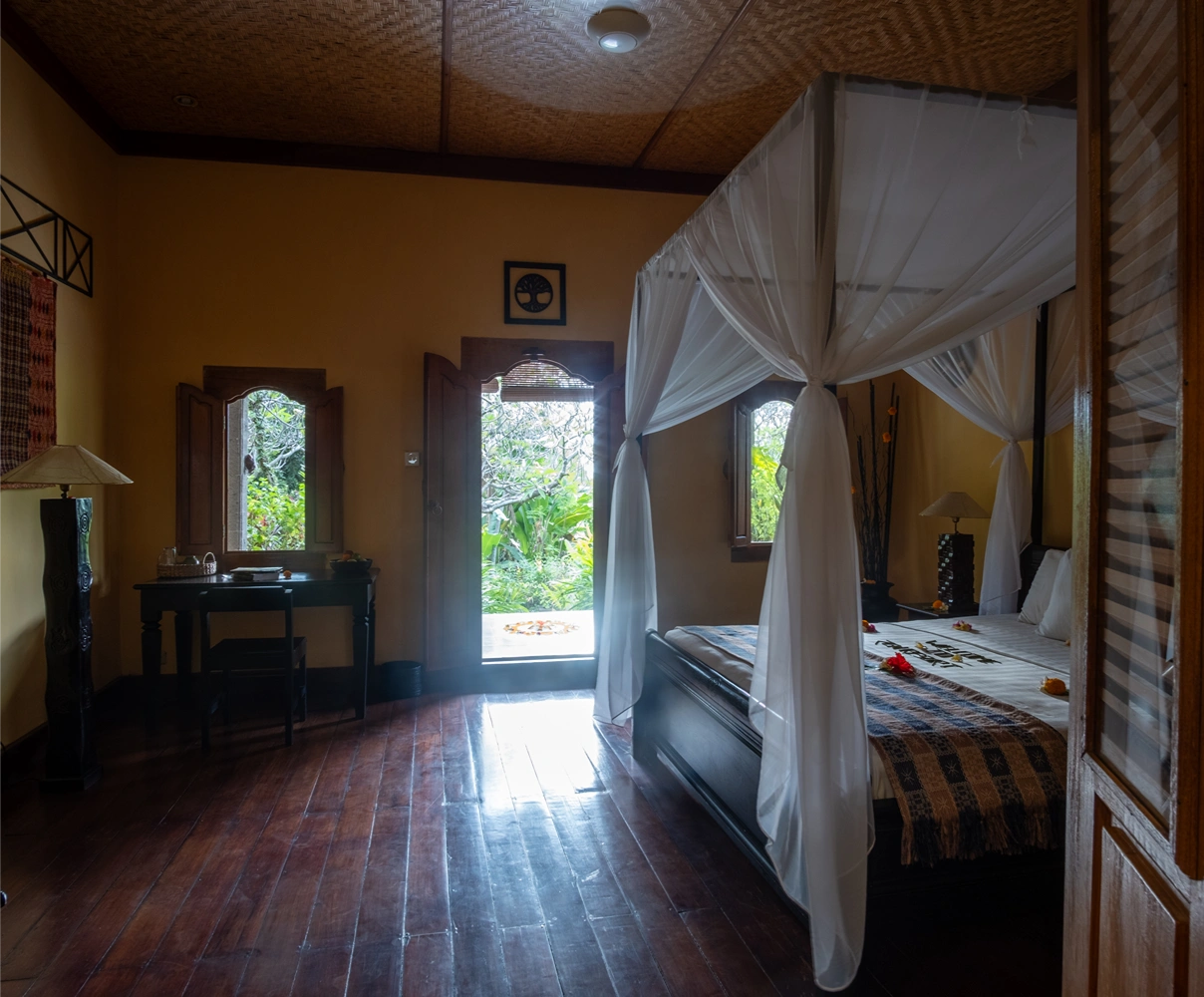 Sunset villa bedroom with a four-poster bed with white curtains and an open door showing green plants outside.