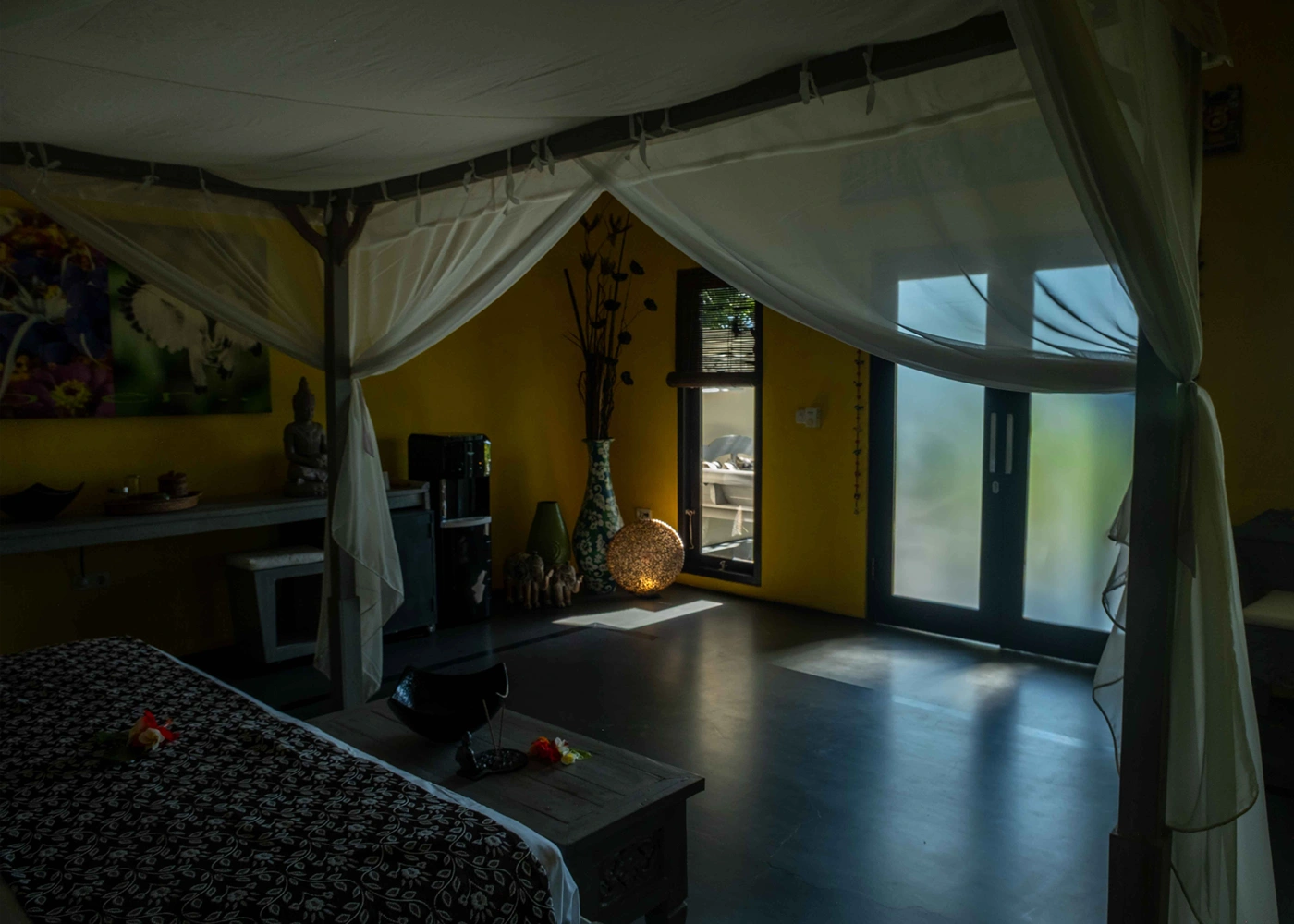 Premium villa with four-poster bed, sheer curtains, yellow walls, decorative vases, and frosted glass door.