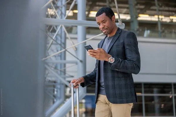 Man in a dark blazer holding a phone and suitcase in an airport terminal.