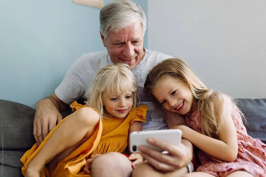 Smiling elderly man sitting on couch with two young girls watching a smartphone together.
