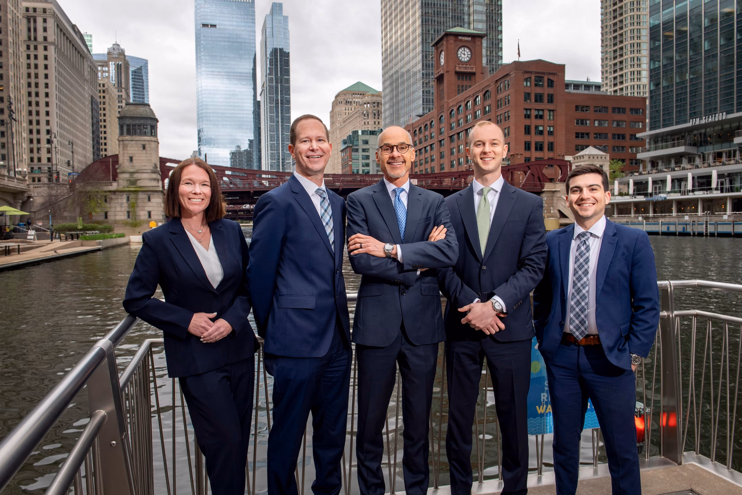 Five business professionals in suits standing on a railing by a river in a city with high-rise buildings.
