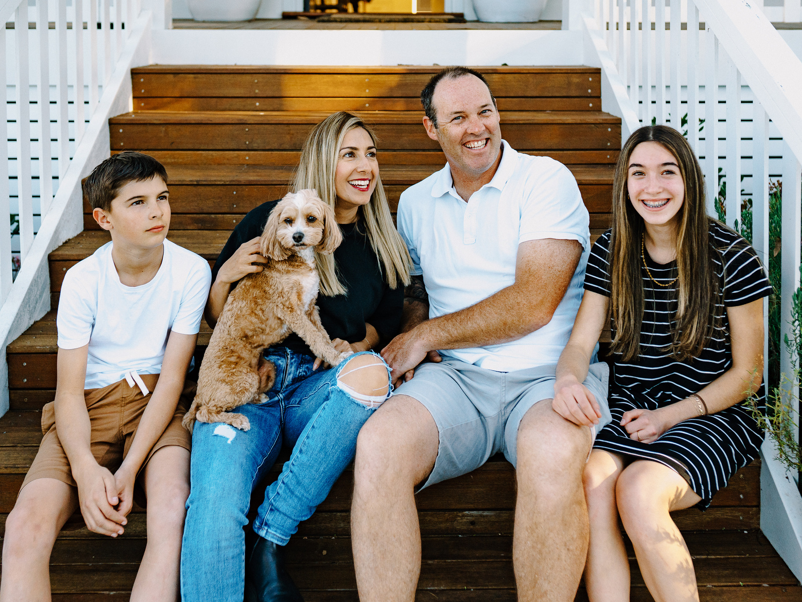 Smiling family of four sitting on wooden steps with a small tan dog on the woman's lap.