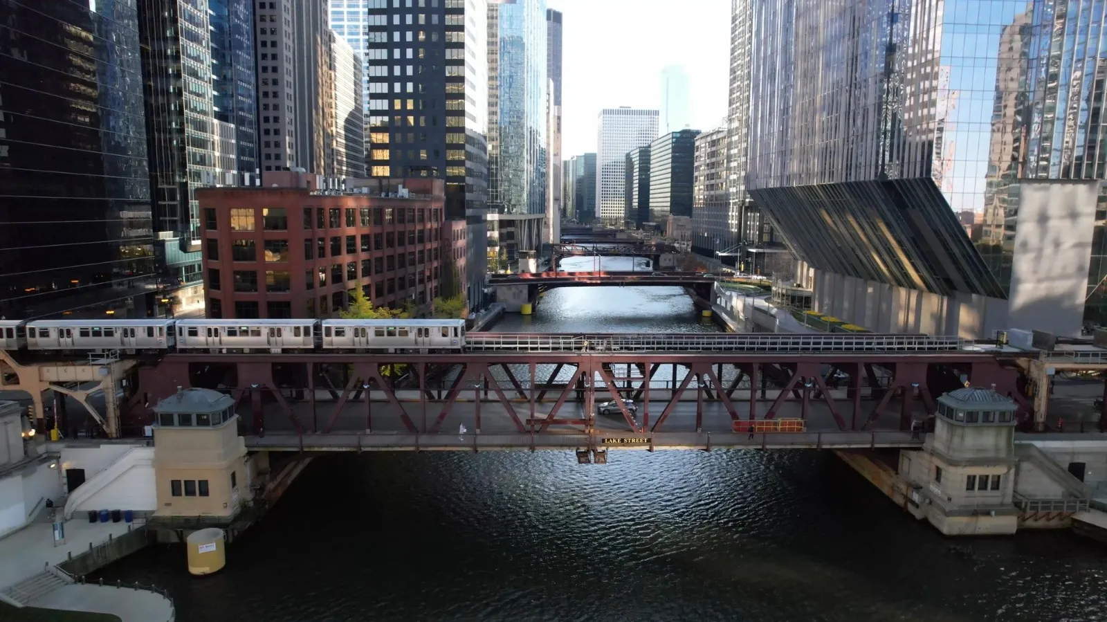 A train crosses a steel bridge over a river in a downtown area with tall glass buildings.