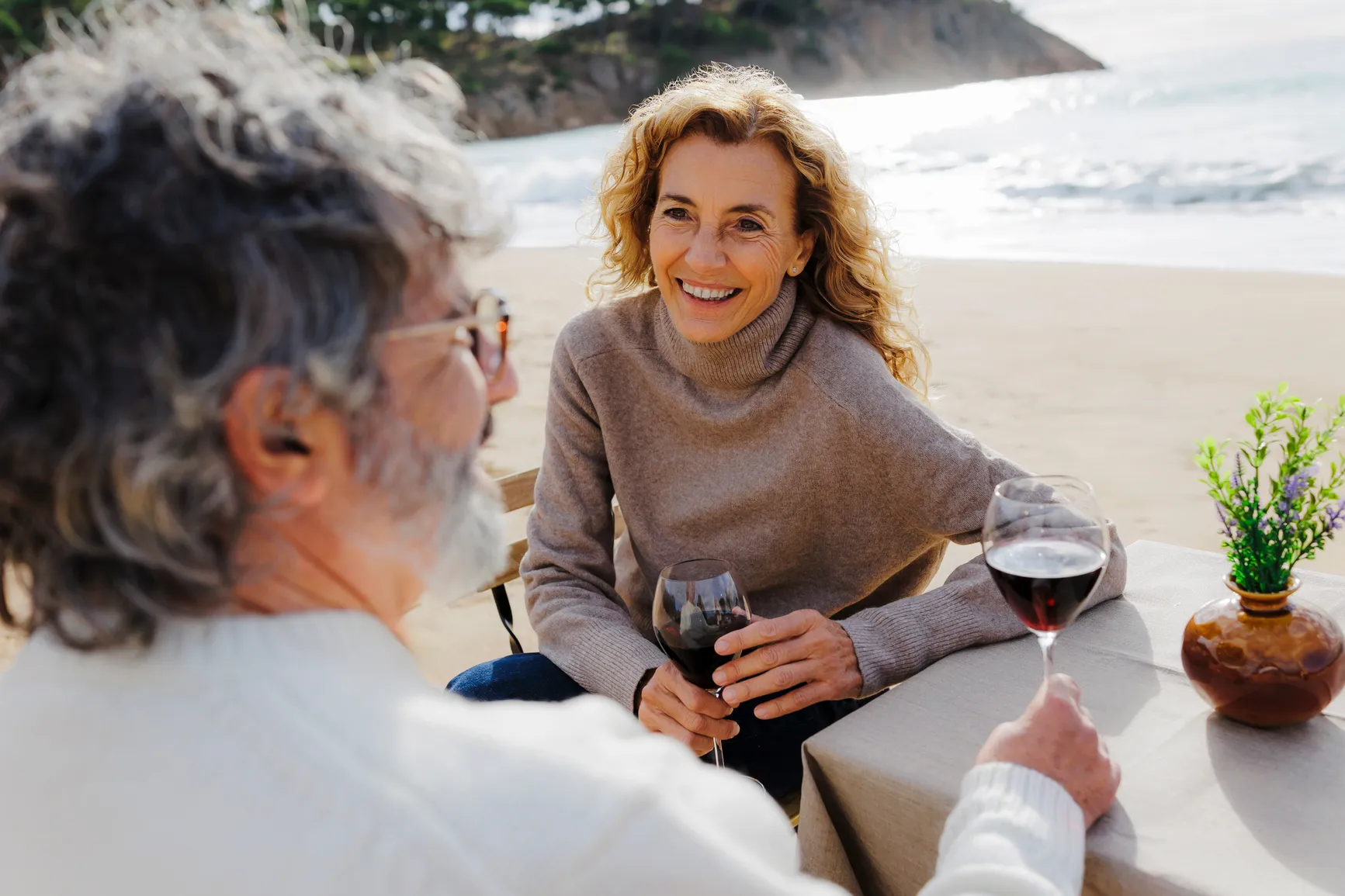 Smiling older couple sitting at a table on the beach, holding glasses of red wine.