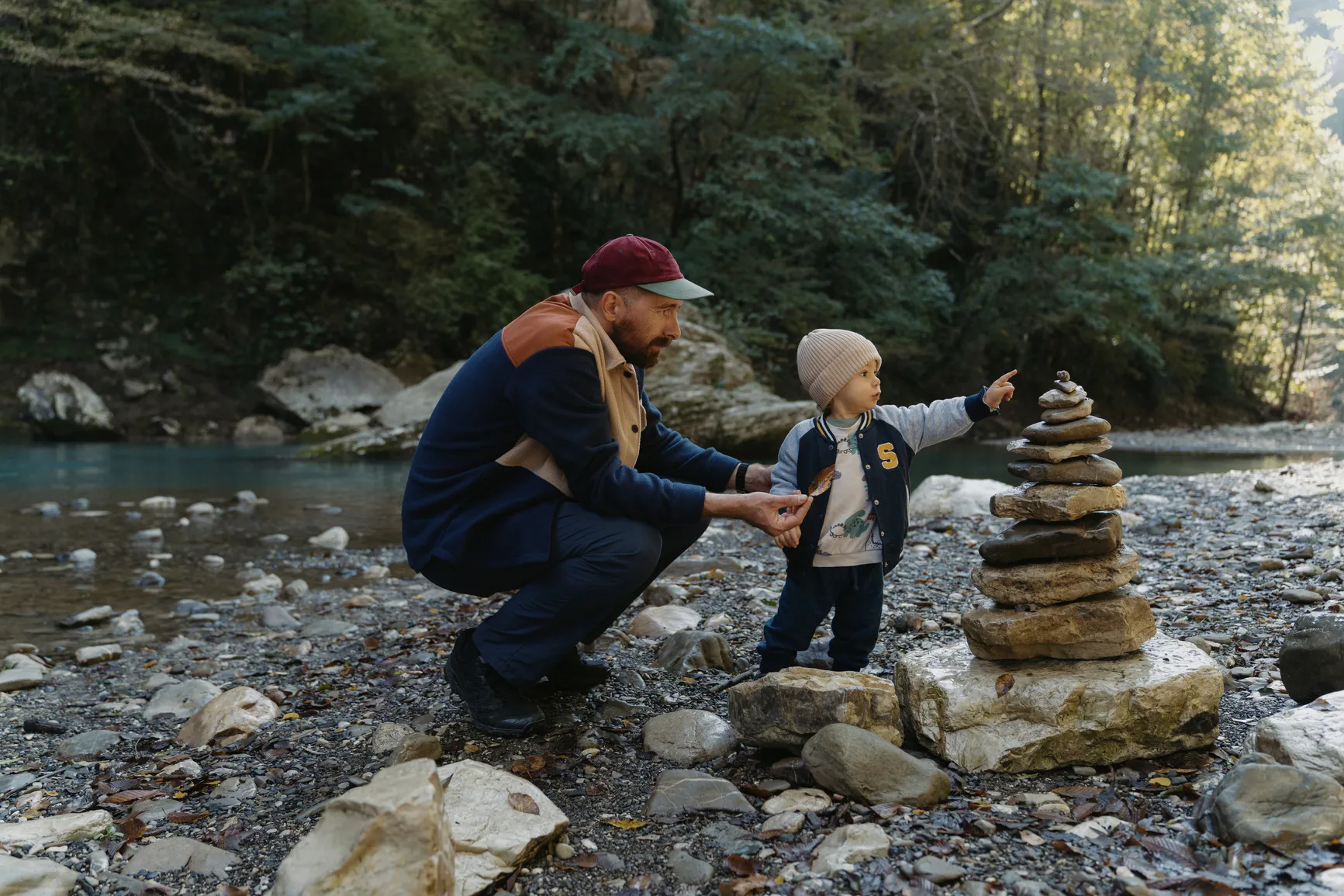 Man crouching by a river holding a leaf while a toddler points at a stacked pile of rocks.