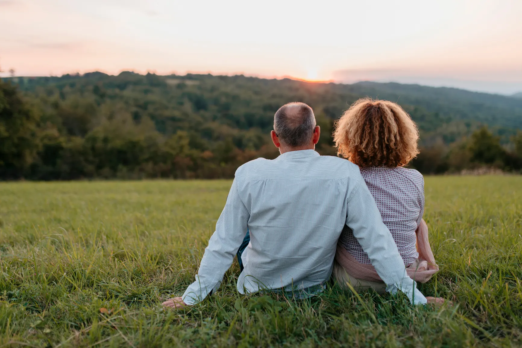Couple sitting on grass, facing a sunset over a forested landscape with hills.