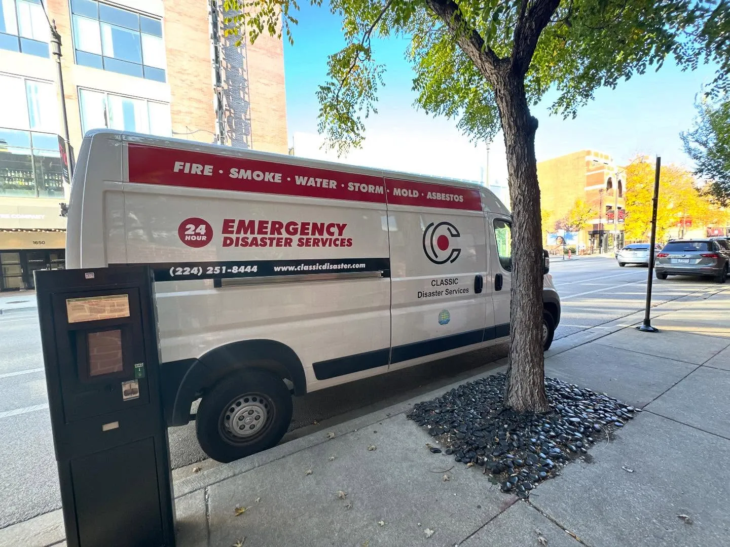 White emergency disaster services van parked by sidewalk with tree and parking meter nearby.