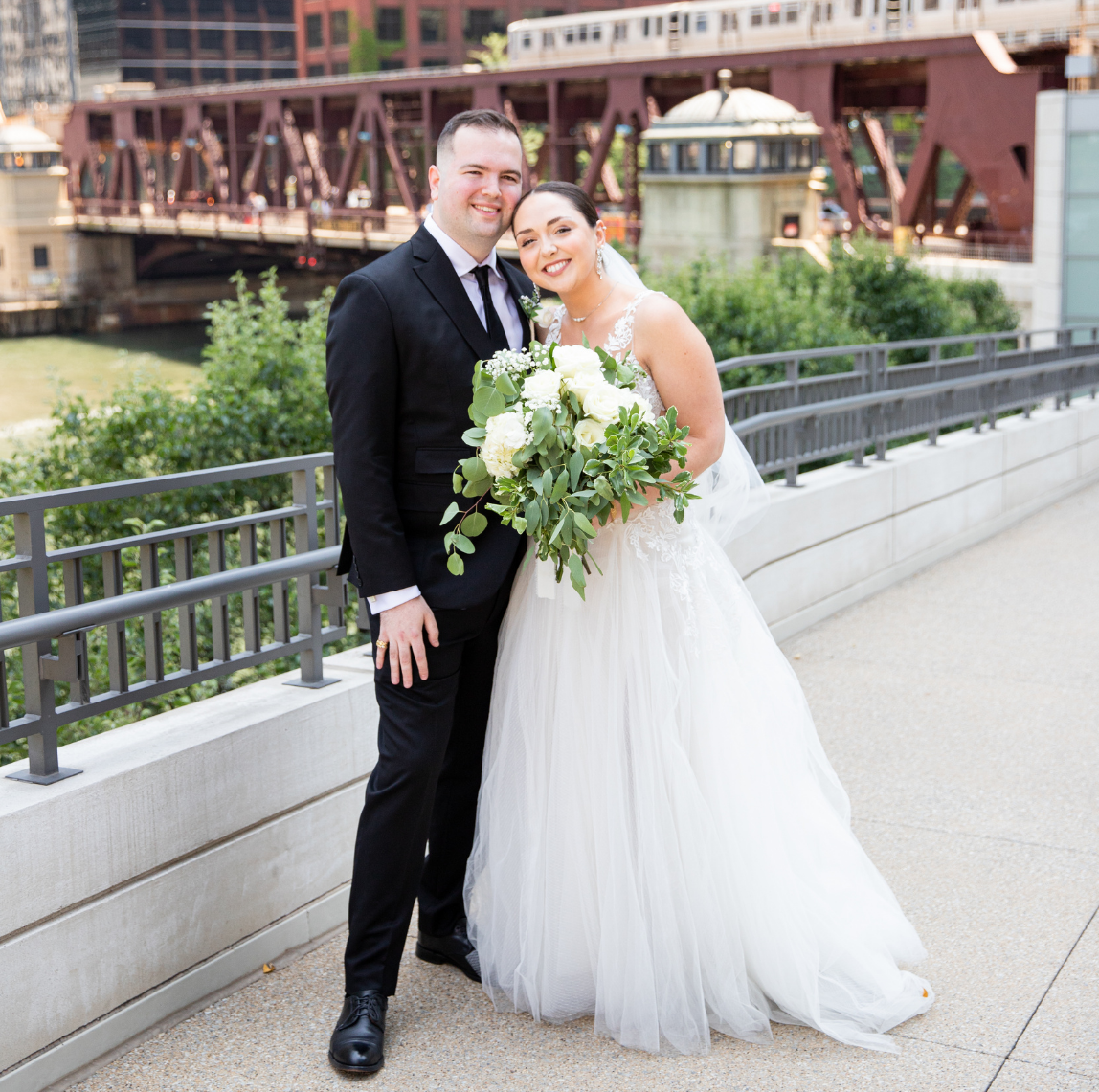 Bride in white dress and groom in black suit hugging outdoors by a river with a bridge behind them.