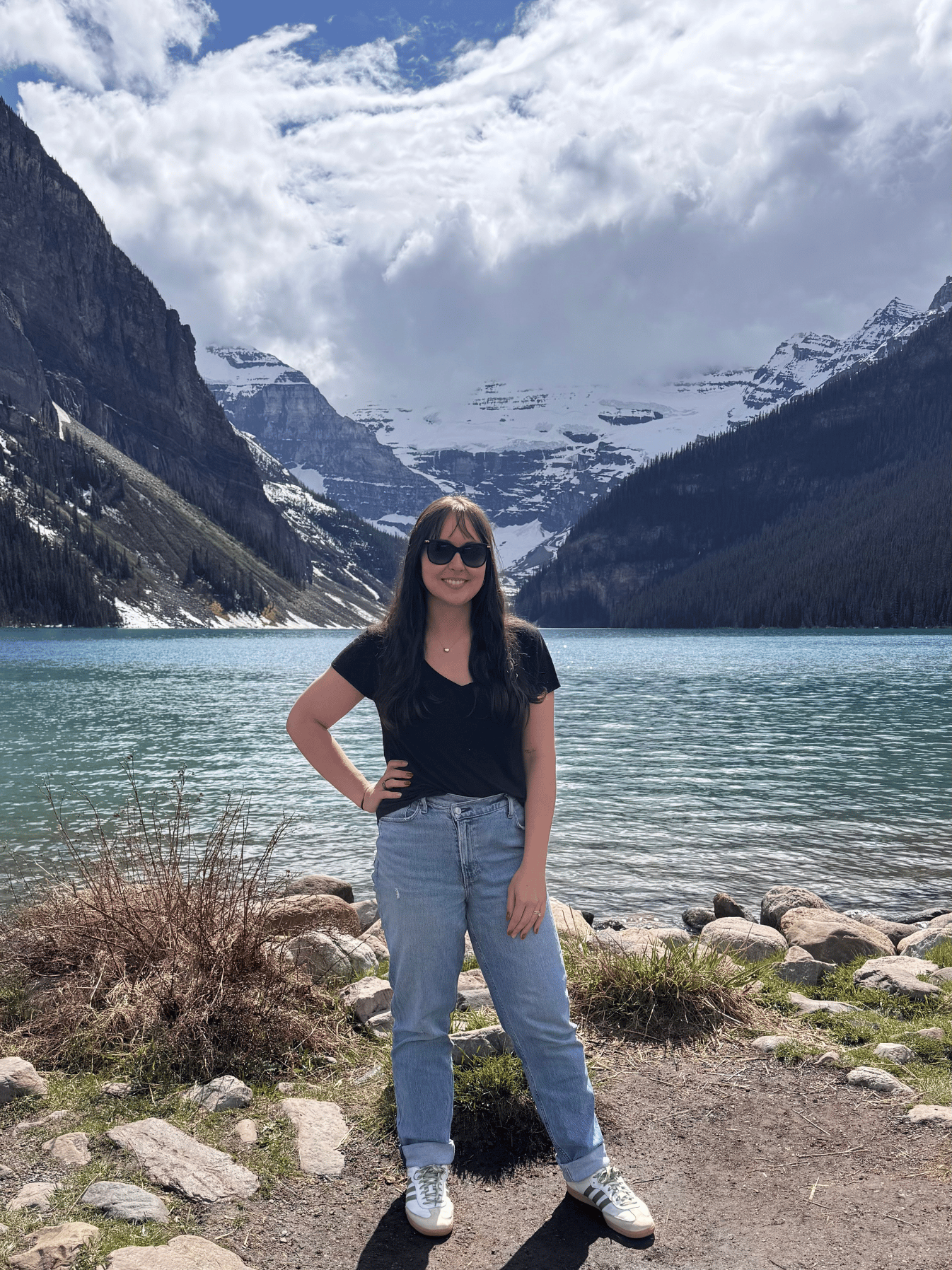 Woman in sunglasses and casual clothes standing by a lake with mountains and snowy peaks in the background.