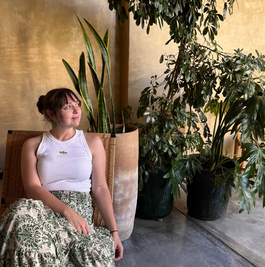 Woman with a white tank top and floral skirt sitting near large potted green plants indoors.