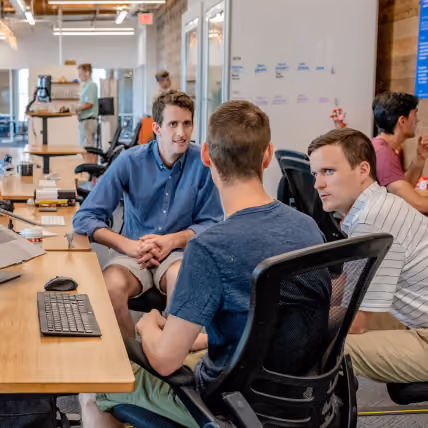 Three men sitting and talking in an office with desks, computers, and a whiteboard.
