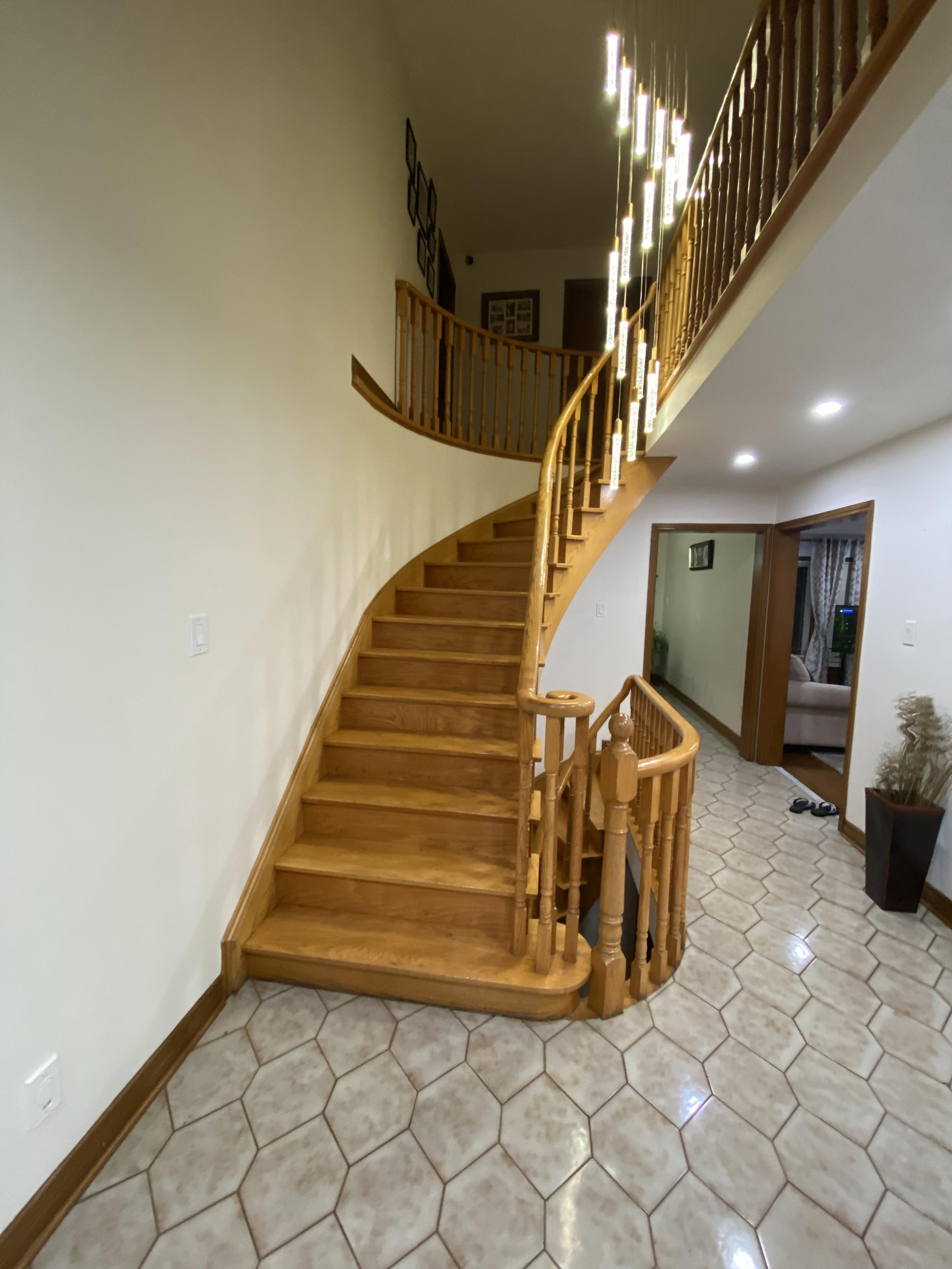 Before photo of a Woodbridge full home renovation showing a builder-grade red oak staircase with light wood stain, wooden spindles, and tiled foyer floor.