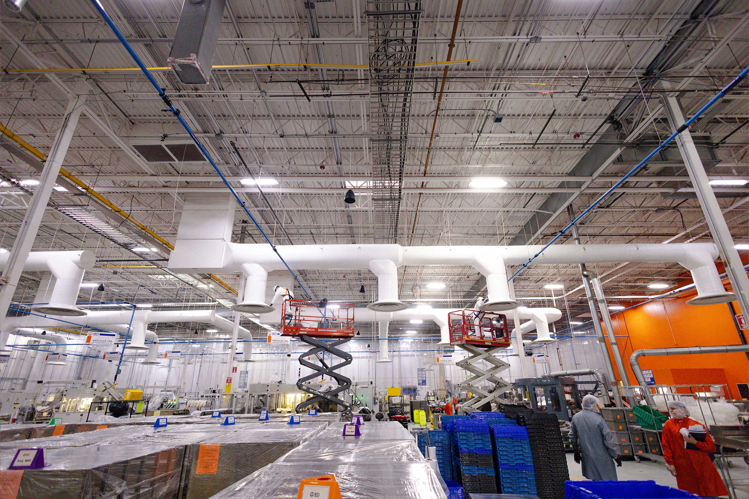 Wide-angle view of a manufacturing facility interior showing overhead ductwork and scissor lifts