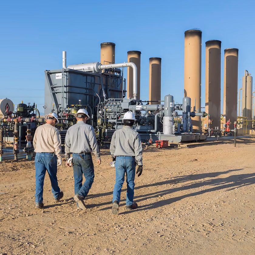 Three workers in helmets walking on dirt toward industrial equipment and tall cylindrical structures.
