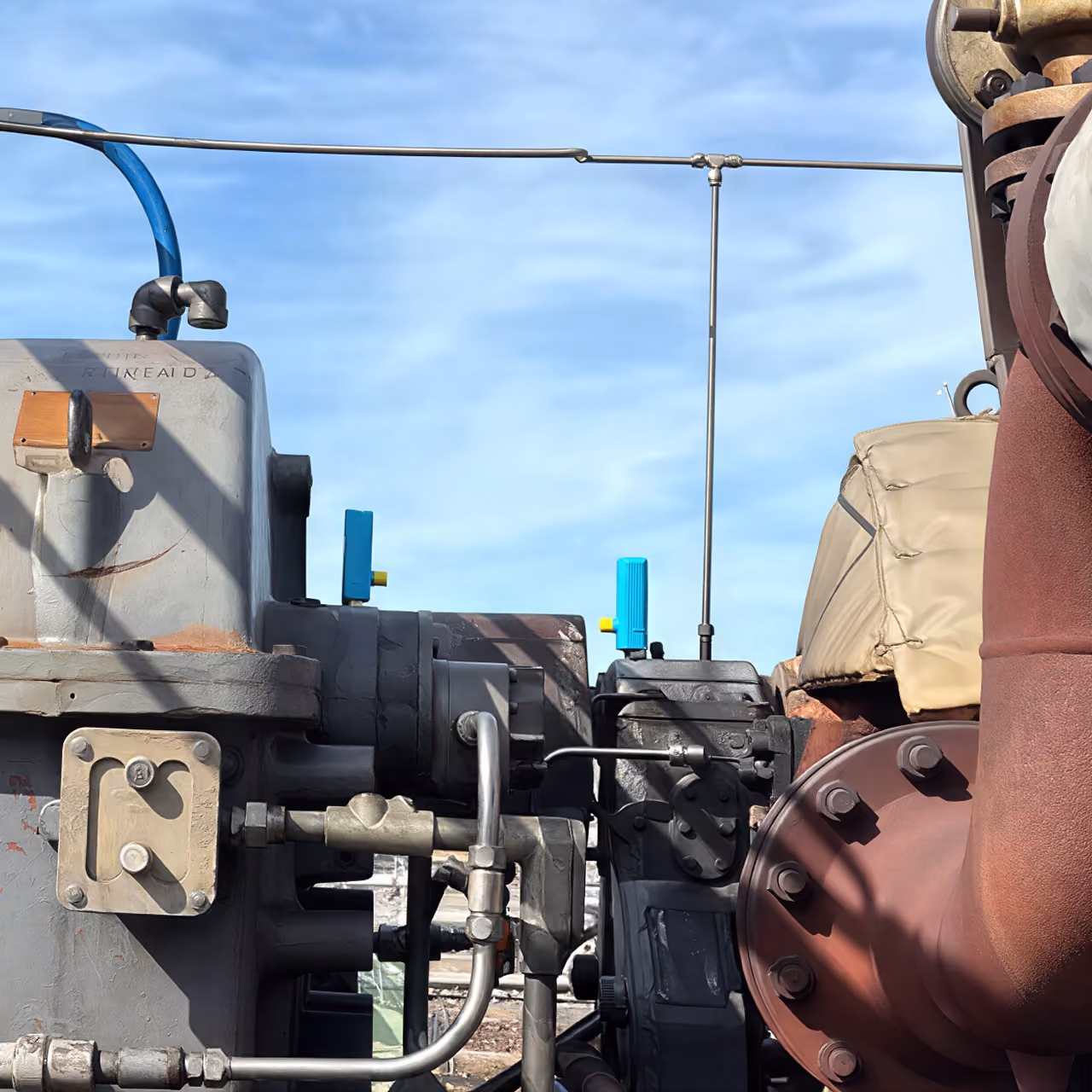 Close-up of industrial machinery parts with pipes, valves, and a blue sky background.