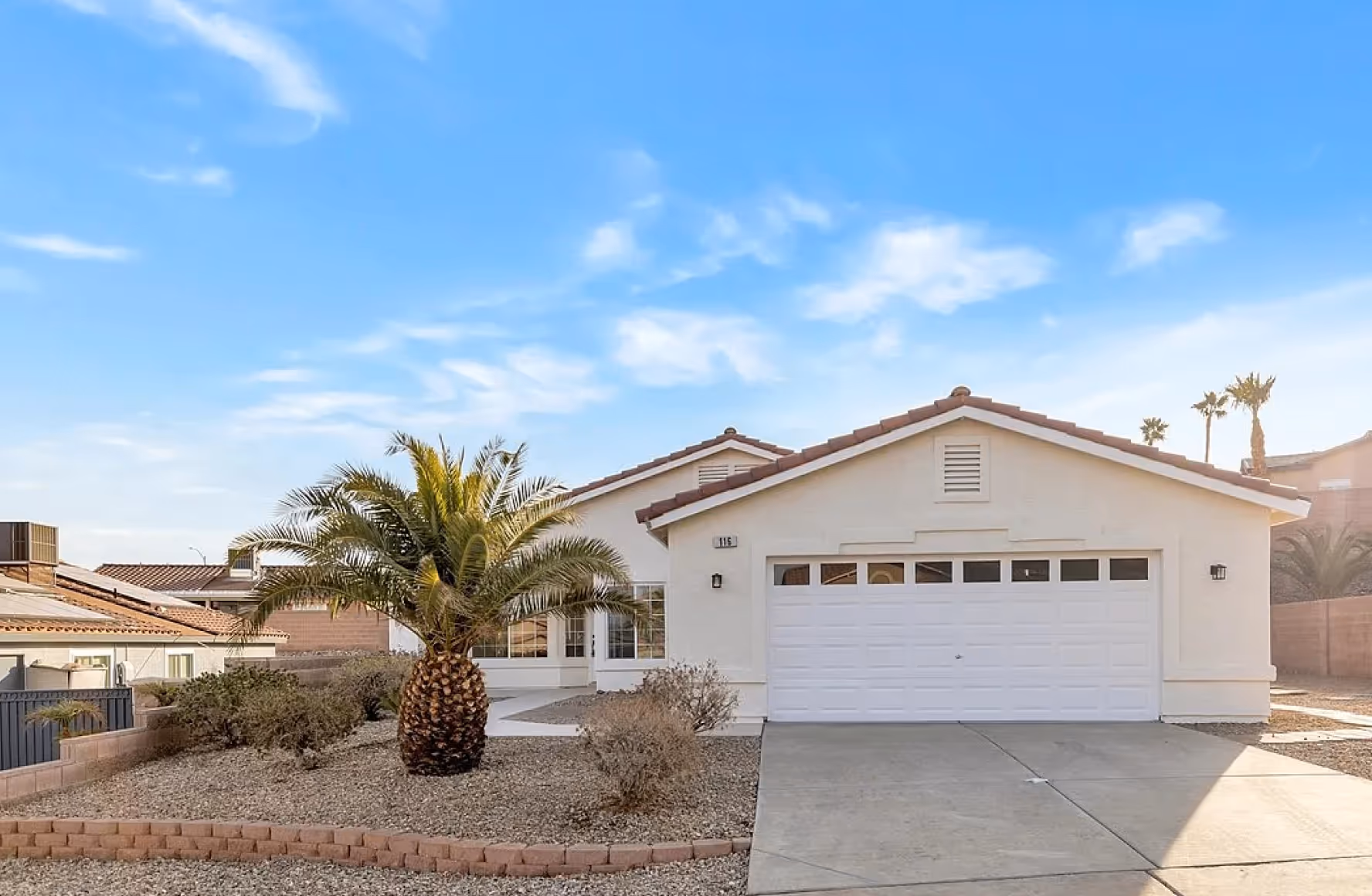 Single-story beige house with a two-car garage, a palm tree, and desert landscaping under a blue sky.