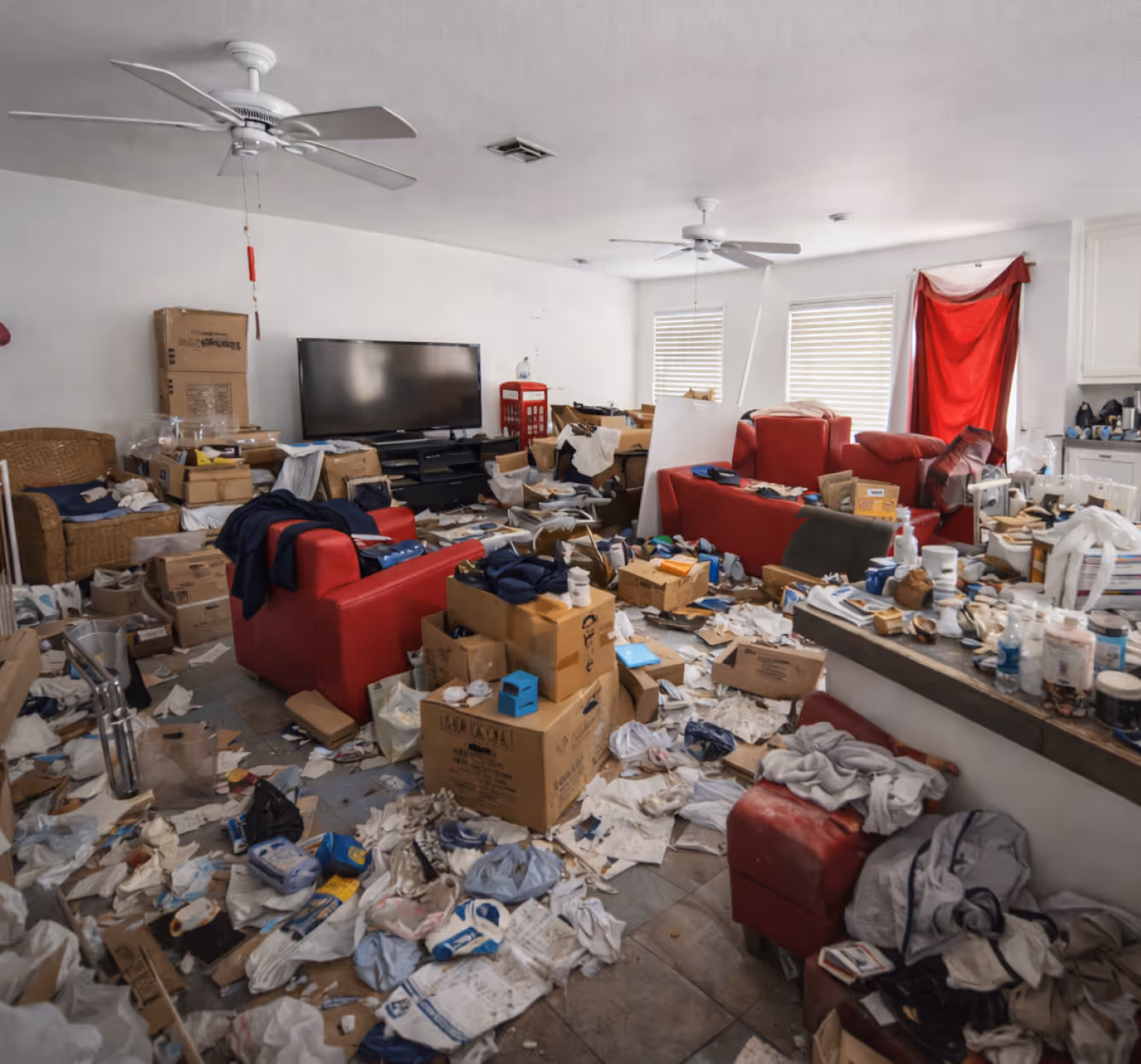 Cluttered living room with red sofas, cardboard boxes, scattered papers, and household items covering the floor and surfaces.