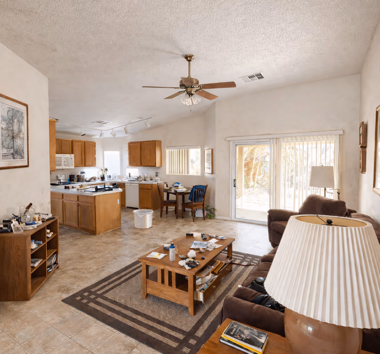 Open-concept living room and kitchen with brown furniture, a coffee table with scattered items, ceiling fan, and sliding glass doors with vertical blinds.