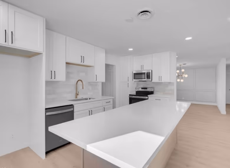 Modern kitchen with white cabinetry, a large white island countertop, stainless steel dishwasher, oven, microwave, and a gold faucet.