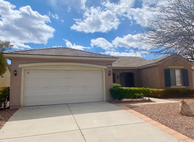 Single-story house with beige exterior, white double garage door, green shrubs by entrance, and partly cloudy blue sky.