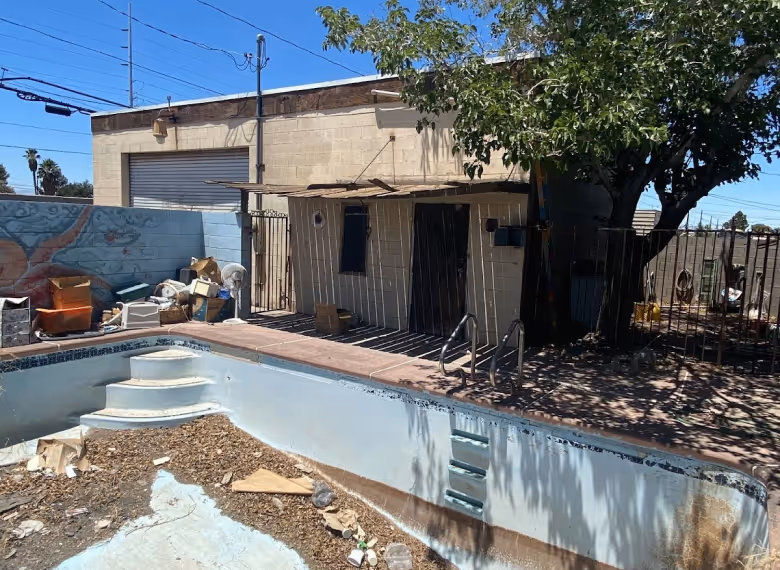 Empty and dirty swimming pool filled with debris in front of a beige building with a garage door and a tree casting shadows.