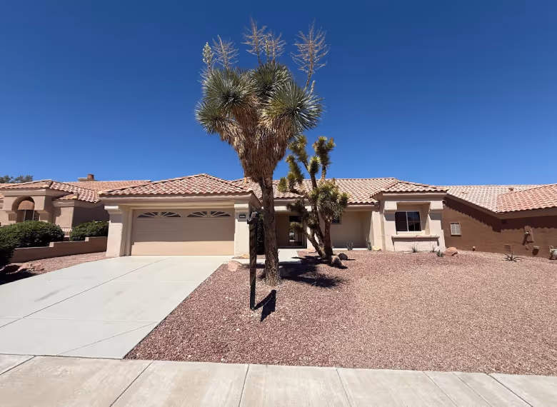 Single-story desert-style house with a tiled roof, a concrete driveway, and drought-tolerant landscaping featuring Joshua trees under a clear blue sky.