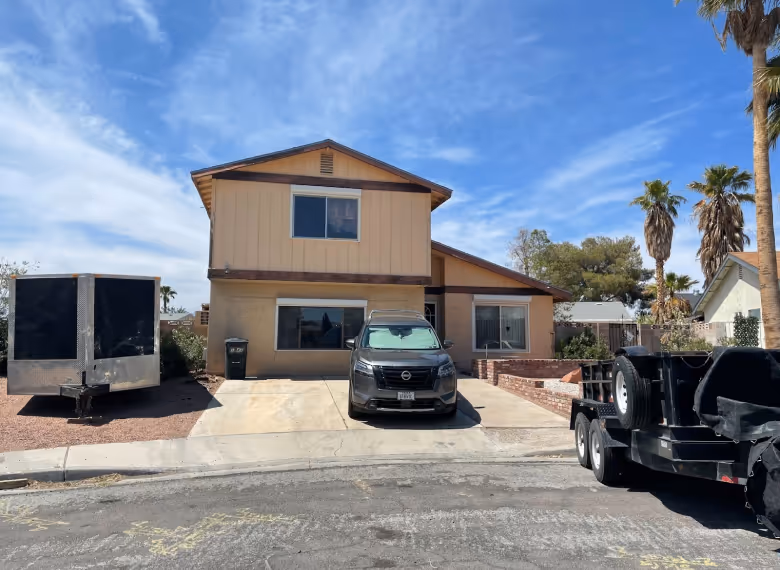 Two-story beige house with a black SUV parked in the driveway and trailers on either side under a partly cloudy sky.