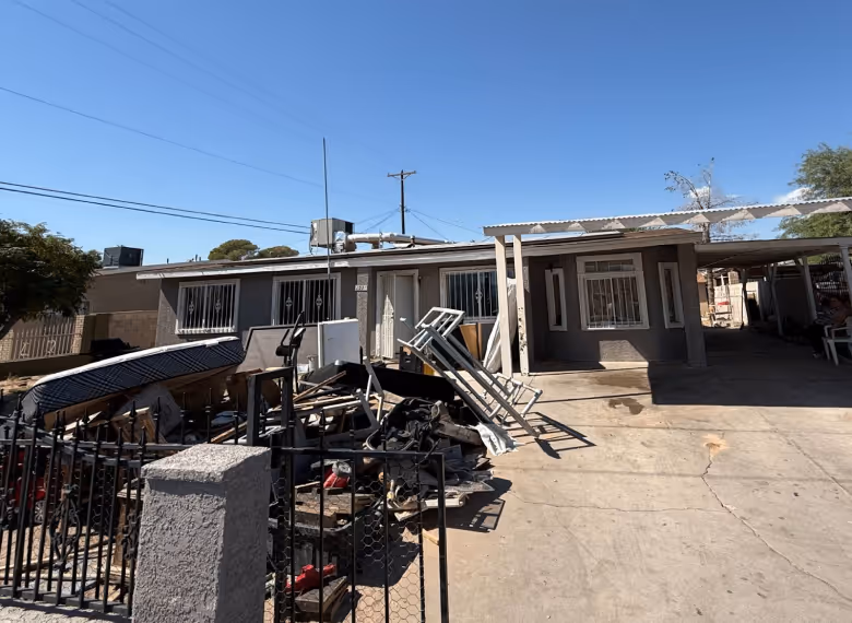 Single-story house with gray exterior and metal bars on windows, cluttered front yard with various discarded items, and concrete driveway under clear blue sky.