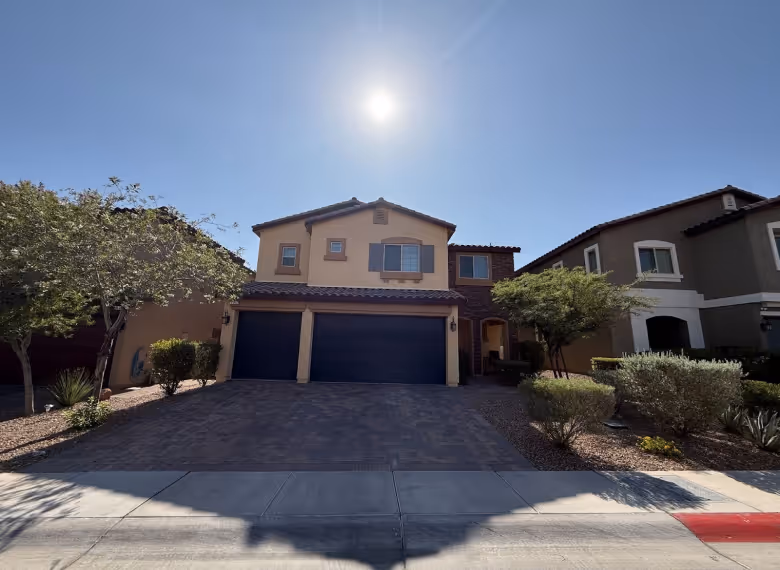 Two-story suburban house with beige and brown exterior, three-car garage, and surrounding desert landscaping under a clear sunny sky.