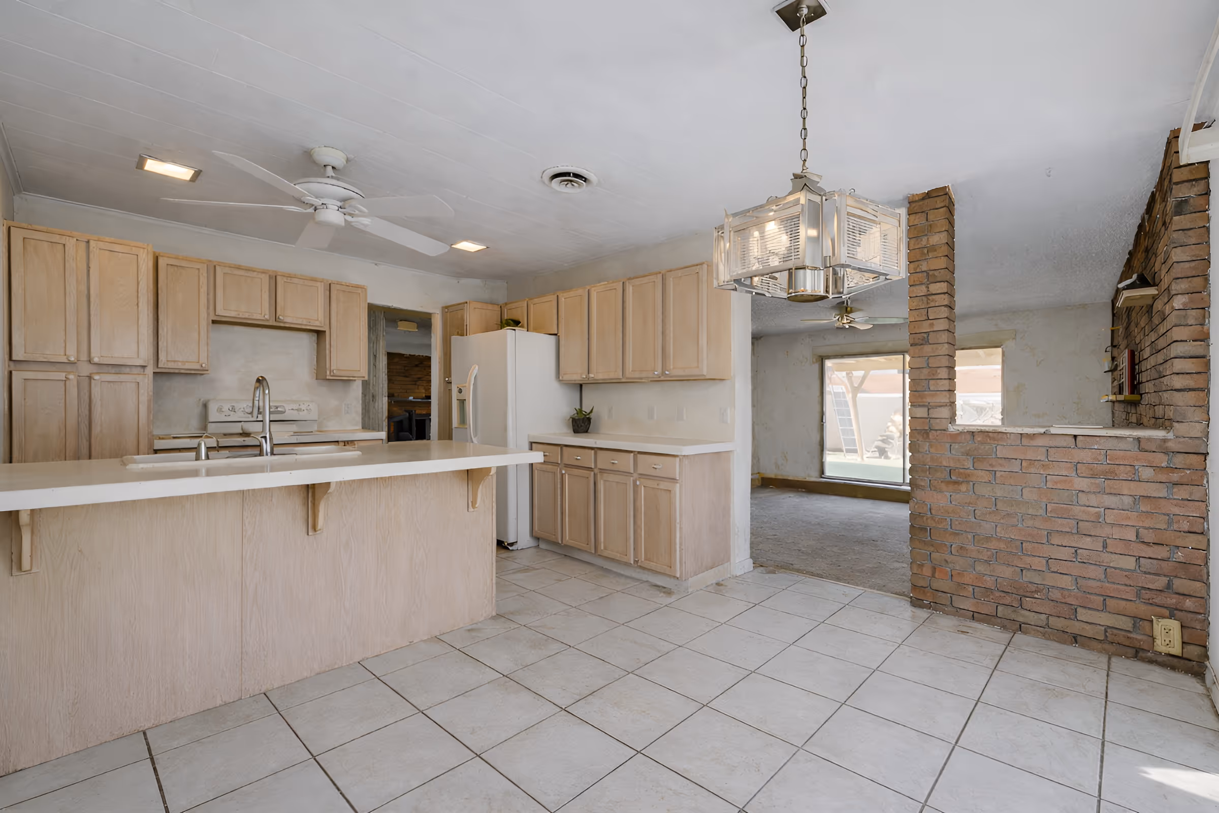 Spacious kitchen with light wood cabinets, white countertops, ceiling fan, and tiled floor next to a dining area with brick partial wall and large window.