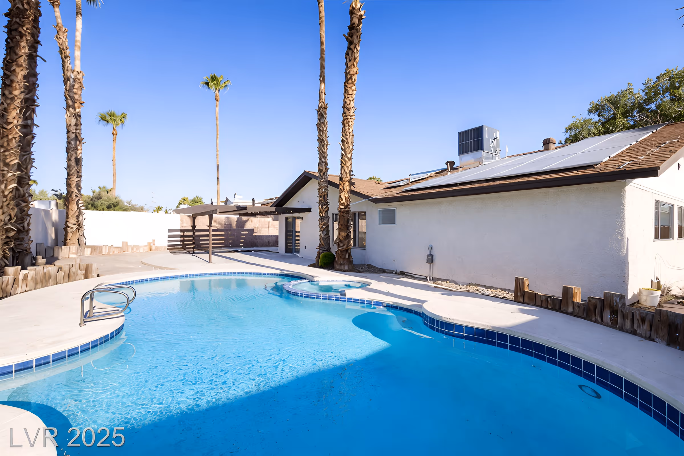 Clear blue swimming pool with white concrete deck and palm trees beside a single-story house with solar panels on the roof.