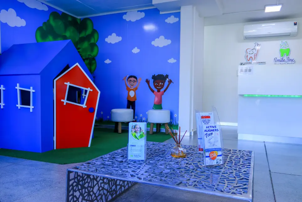 Bright dental clinic waiting area with a blue playhouse, cartoon kids mural, and a metal table with pamphlets.