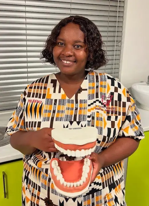 Smiling woman in patterned dental uniform holding a large model of teeth and gums for education.