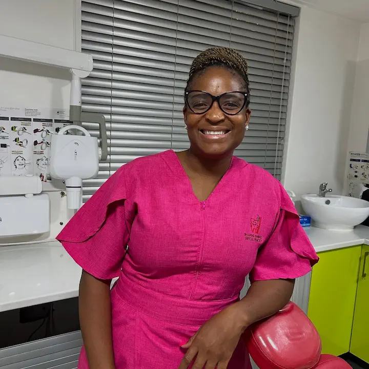 Smiling female dentist in pink scrubs and glasses leaning on a red dental chair in a clinic.
