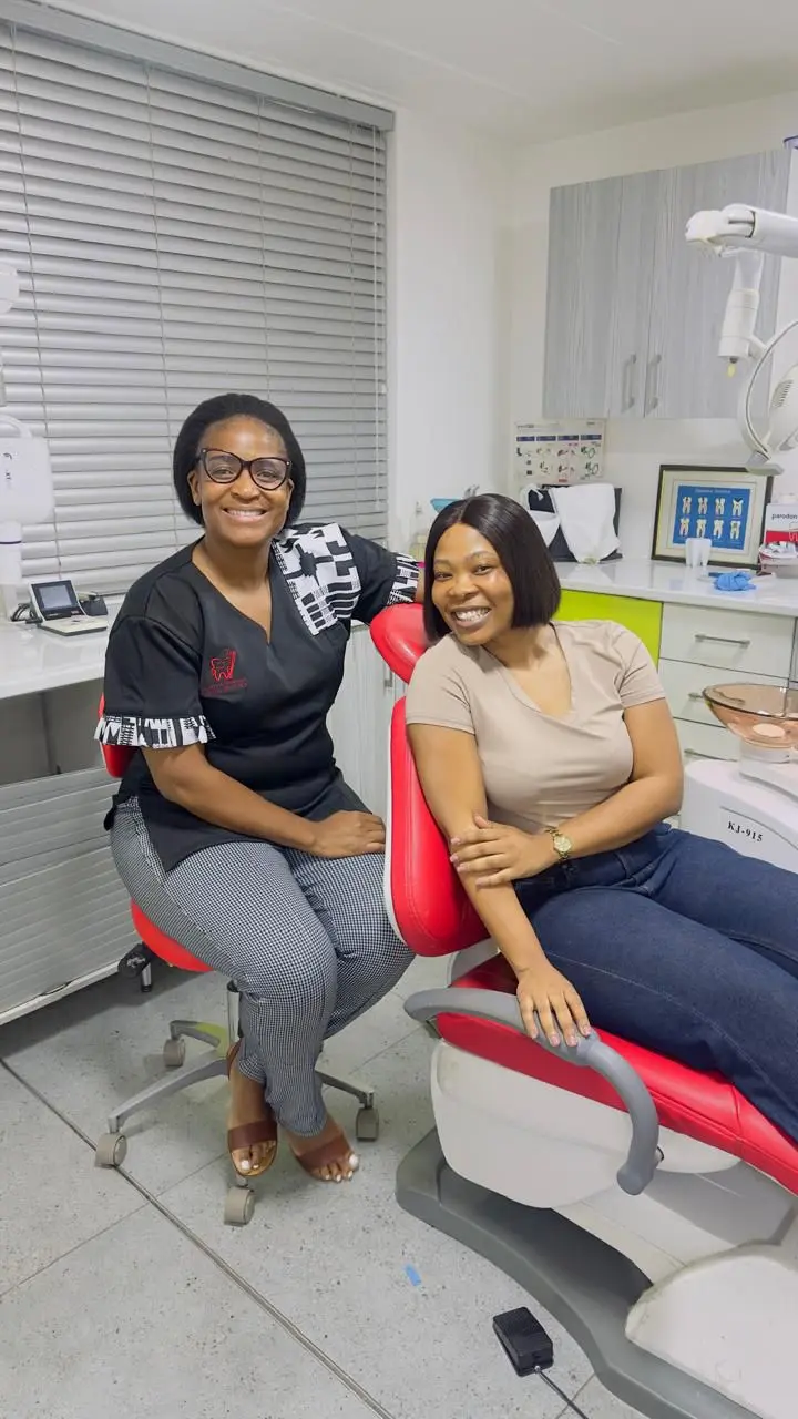 Two smiling women in a dental clinic, one in a dental chair, the other in professional attire beside her.