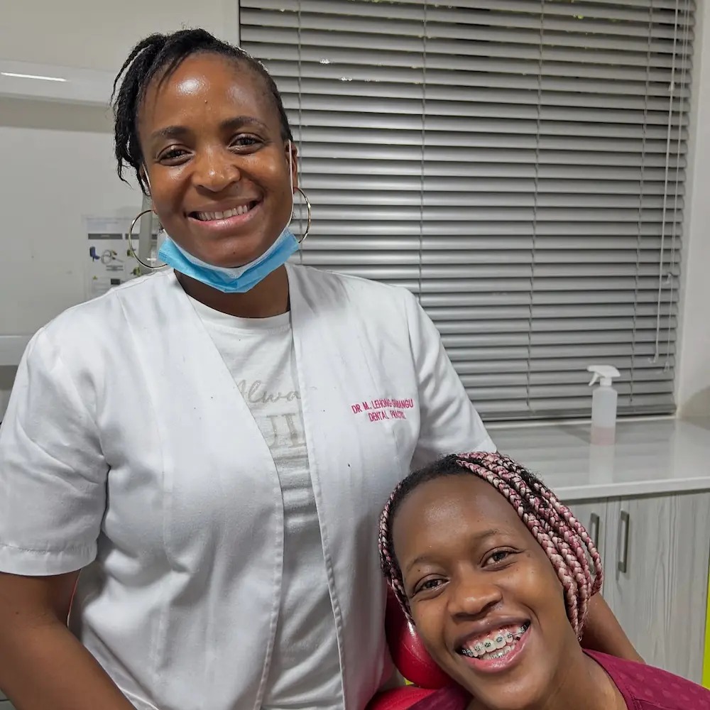 Smiling female dentist with mask below chin stands next to a happy patient with braces in dental clinic.