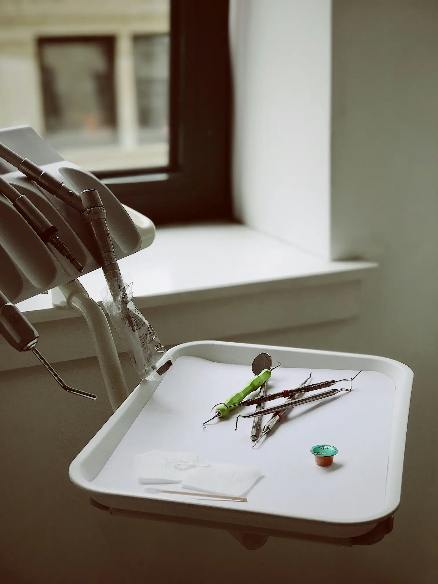 Dental tools and instruments arranged on a white tray near dental equipment by a window.