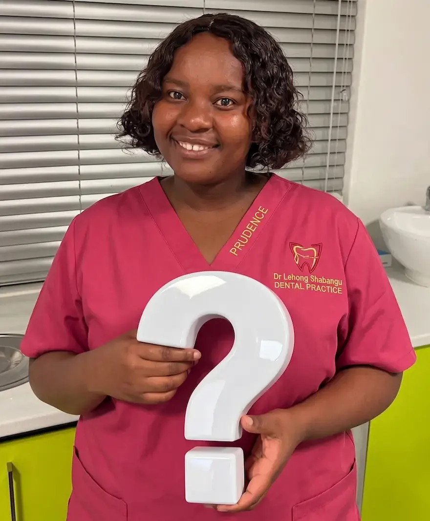 Smiling dental nurse in red scrubs holding a large white question mark in a dental clinic.