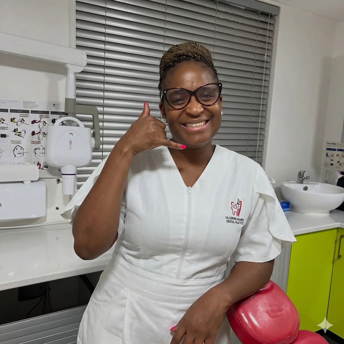 Smiling dental professional in white uniform gesturing phone sign in a clinic room with dental equipment.