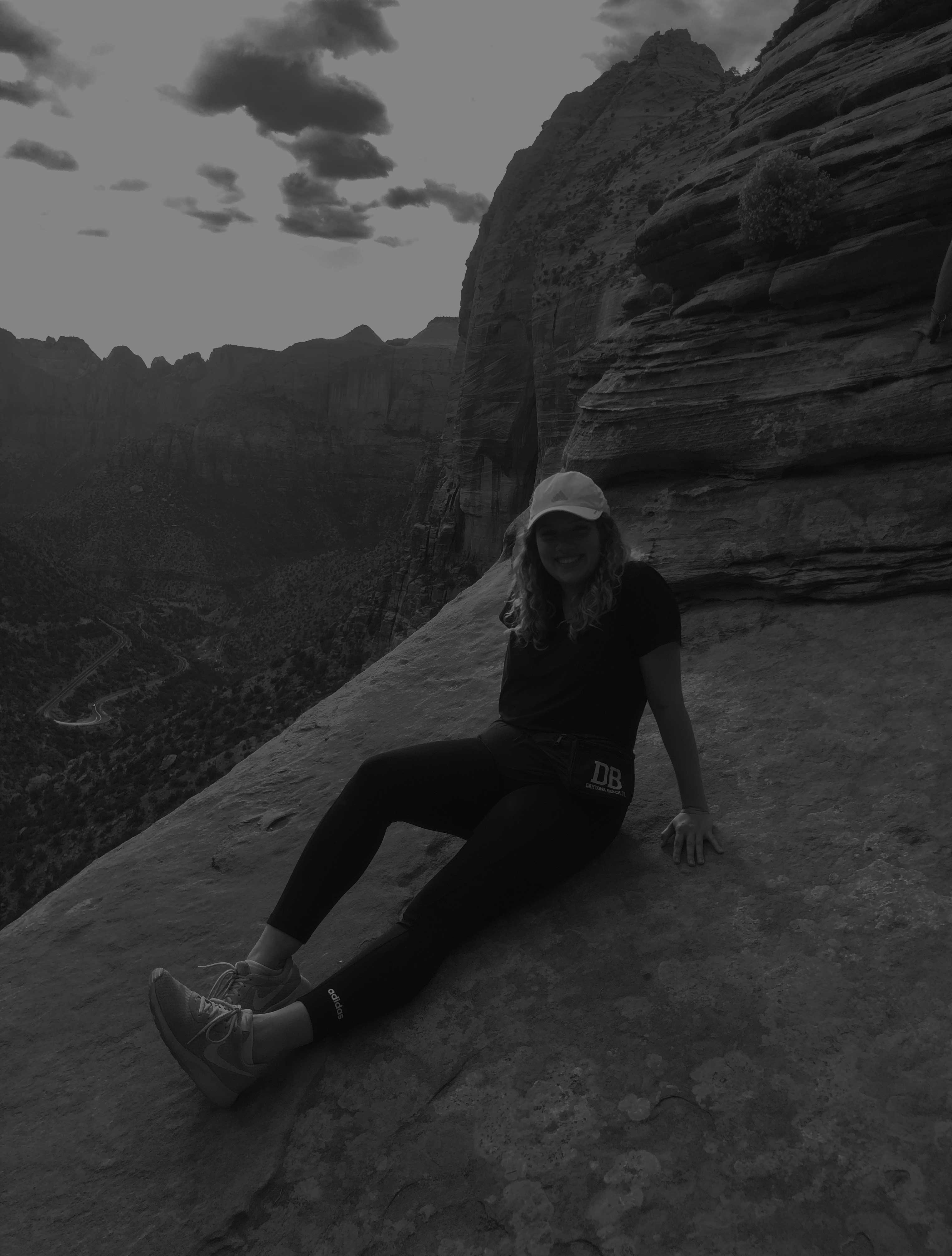 Woman in cap and athletic wear sitting on a rock ledge with mountainous landscape in the background.