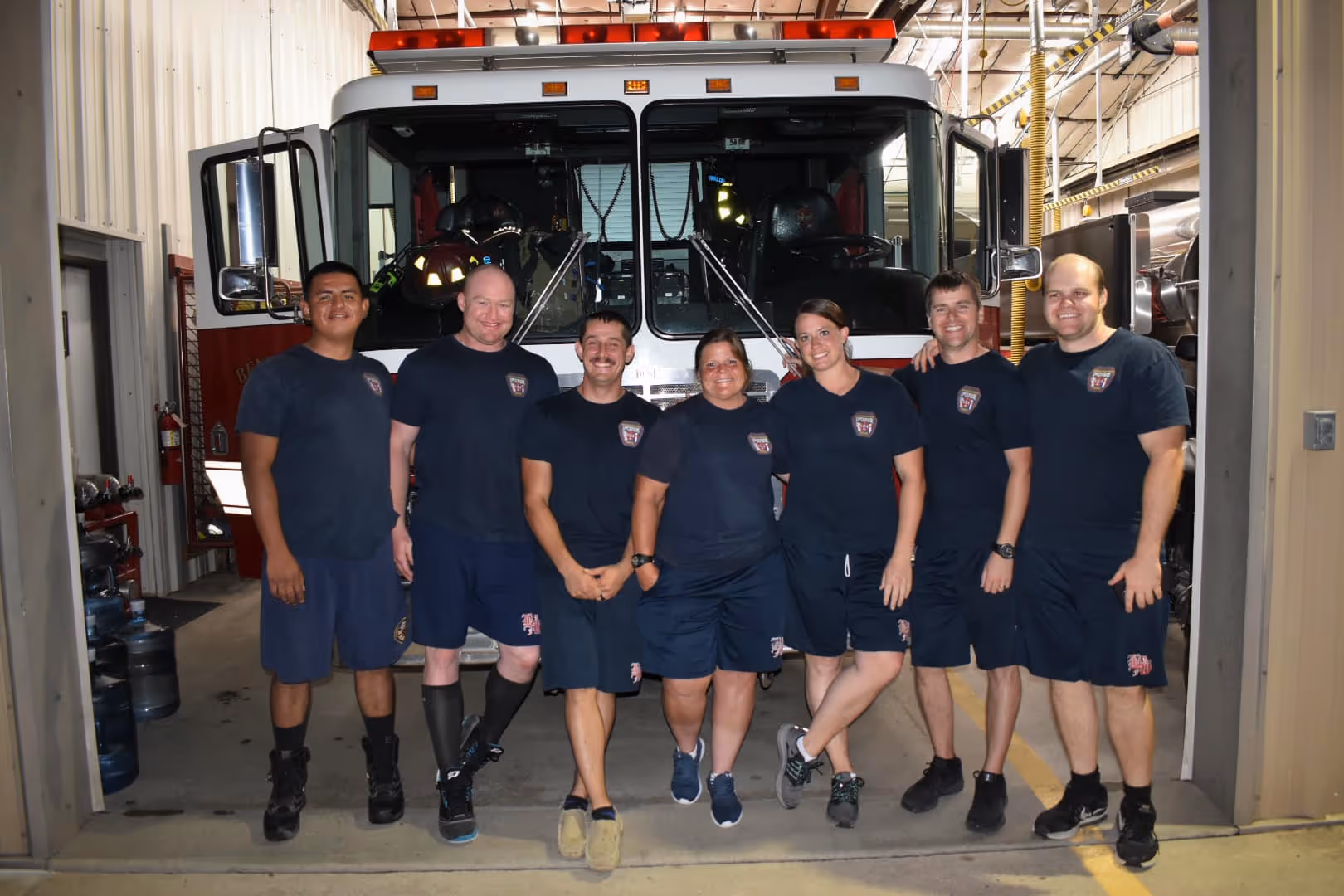 Seven firefighters in blue shorts and t-shirts standing in front of a fire truck inside a station.