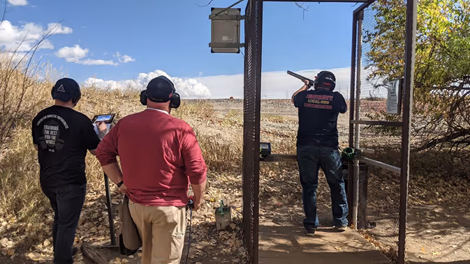 Three men at an outdoor shooting range, one firing a shotgun at a target downrange.