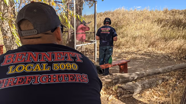 Two men wearing firefighter shirts near a shooting range with targets in a dry, grassy area.