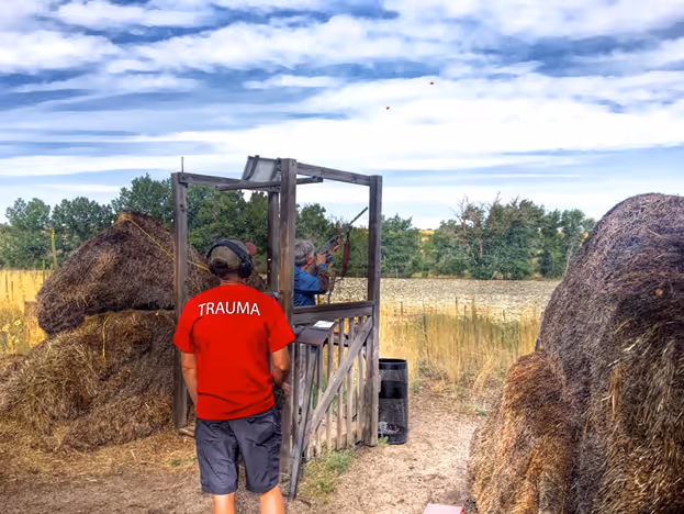 Two people at an outdoor shooting range, one aiming a shotgun, another in a red shirt labeled TRAUMA.