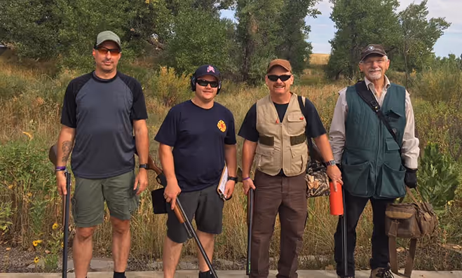 Four men standing outdoors holding shotguns, dressed casually for shooting or hunting.