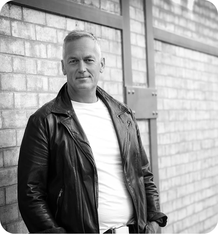 Middle-aged man in a leather jacket and white shirt leaning against a brick wall.