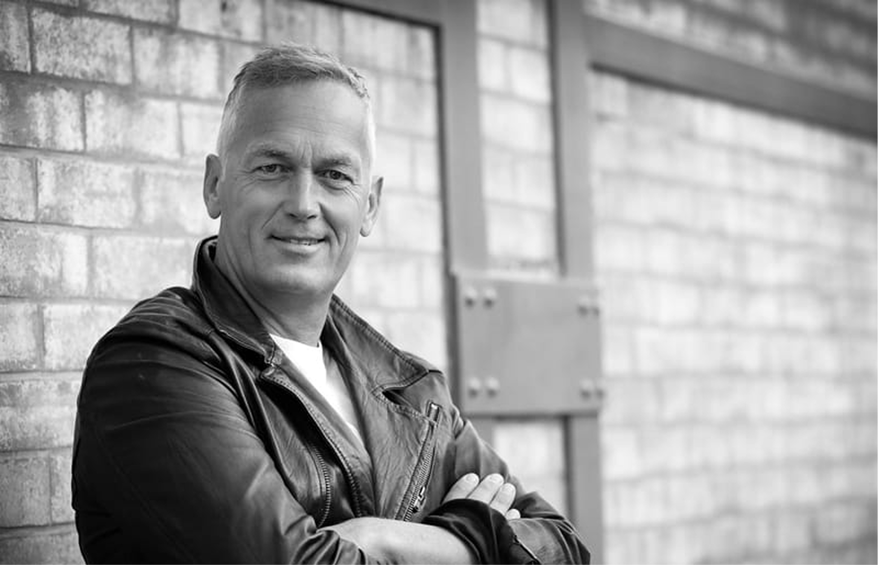 Smiling middle-aged man in a leather jacket standing with arms crossed against a brick wall.